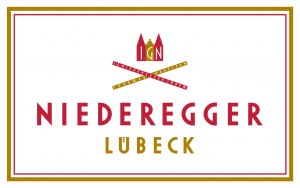Das Logo von Niederegger Lübeck besteht aus rotem und goldenem Text mit einer stilisierten Krone darüber und zwei goldenen und roten Bannern, die sich oben kreuzen, alles innerhalb eines umrandeten weißen Rechtecks.